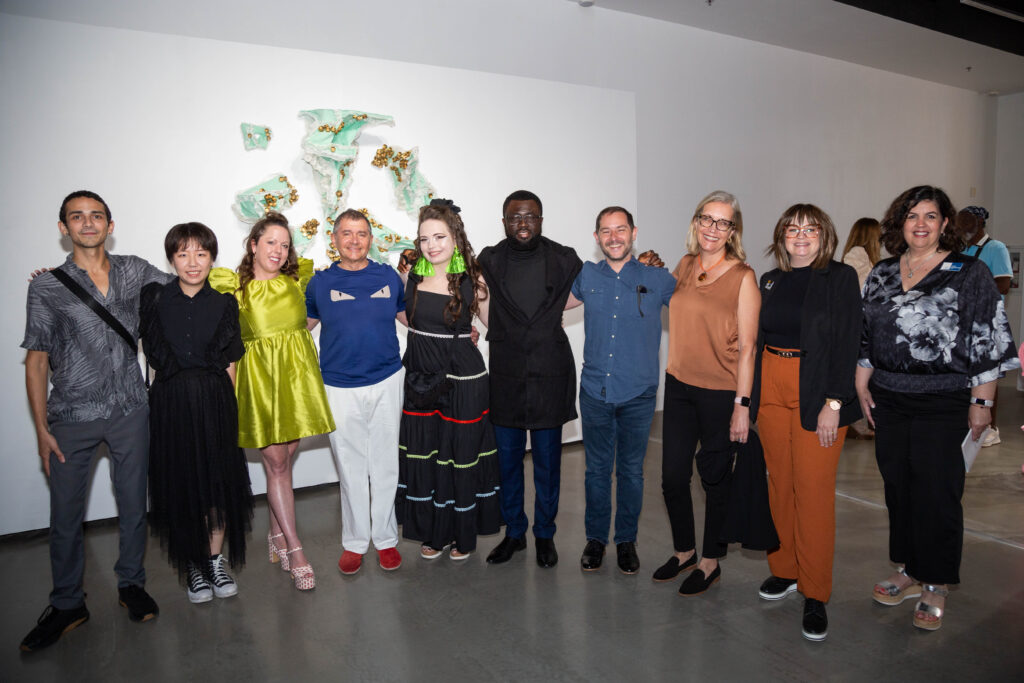 Group of ten people standing side by side in a gallery space, smiling at the camera.