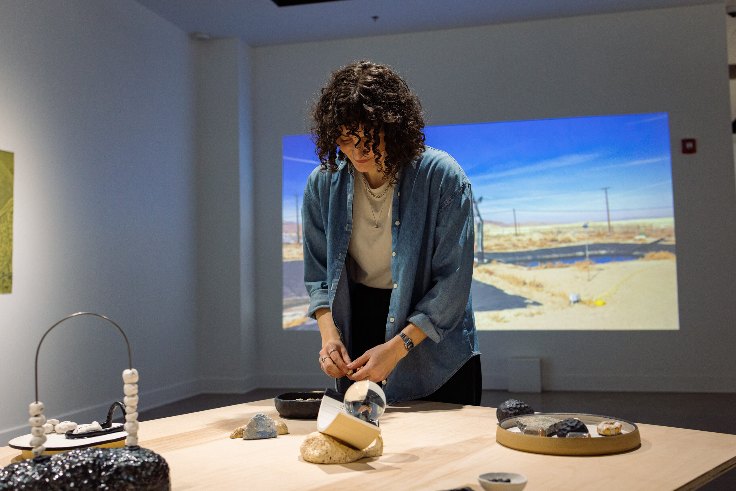 Artist Amanda Leigh Evans arranges objects on a tabletop installation in a gallery, with a projected desert landscape visible in the background.