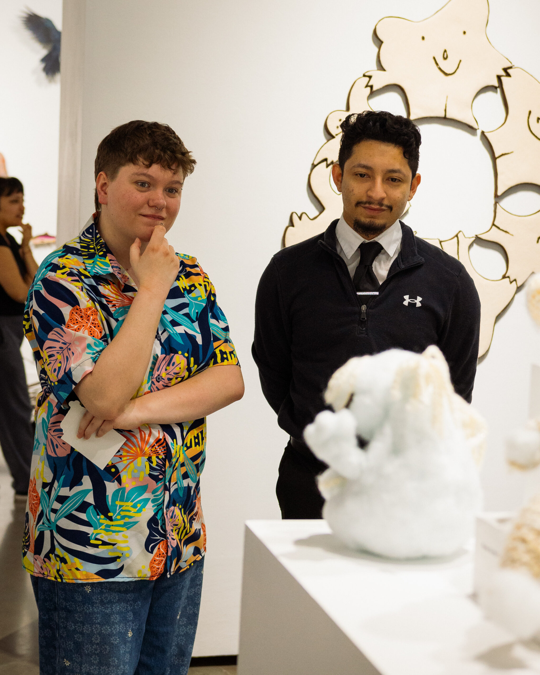 Two visitors stand in an art gallery looking at a white sculpture displayed on a pedestal.