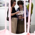 two people reading an open exhibition catalog behind a pink sculputre