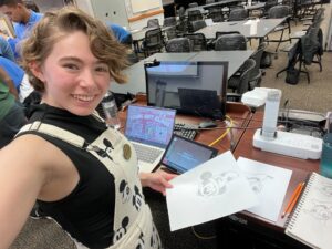 A selfie of Hailey behind the lecturer's desk in a classroom. On one side of the desk is a laptop with a screen reading "Toon Art Class." On the other side of the desk, a document camera displays pencil drawings of Disney cartoon characters.