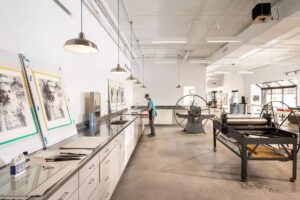A spacious printing studio with large equipment and an artist standing by a workbench.