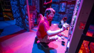 A student wearing safety goggles kneels while painting a colorful themed set, surrounded by props and textured walls under vibrant stage lighting.