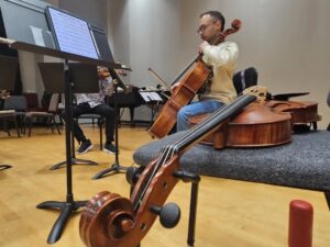 Two musicians rehearse in a music room, one playing cello and the other playing violin.
