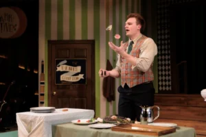 Actor performs onstage during UCF Theatre's production of One Man, Two Guvnors, tossing food in the air beside a dining table set with plates and serving dishes.