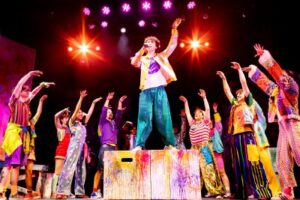 Solo singer on colorful theatre stage with ensemble.