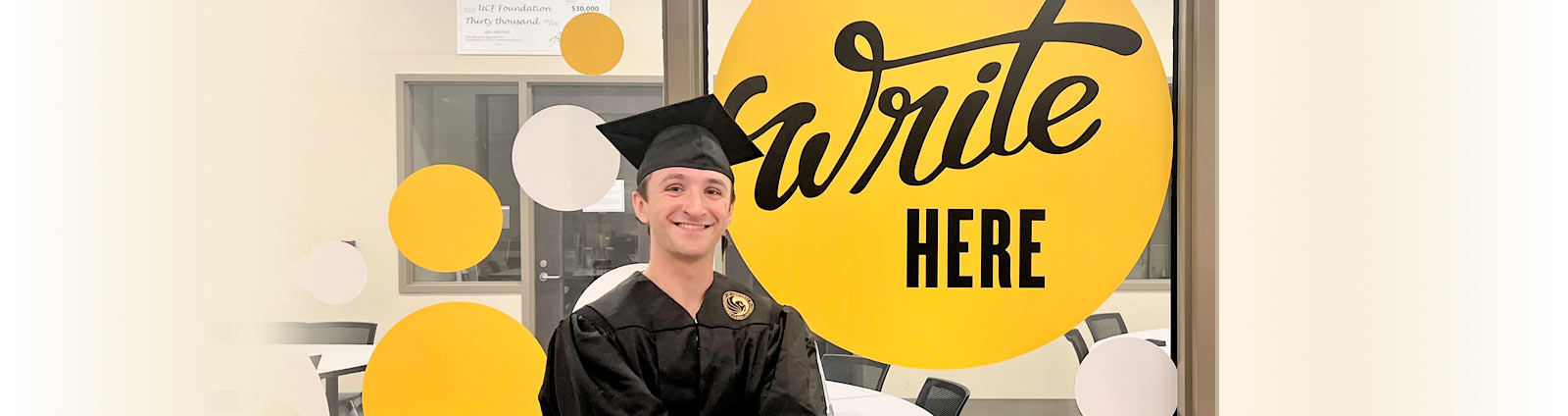 Student in graduate cap and gown stands outside University Writing Center