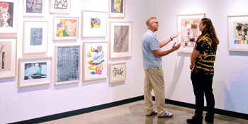 Two people converse in front of a wall of framed artworks