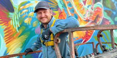 Christian Stanley poses on a lift in front of a mural wearing painting coveralls