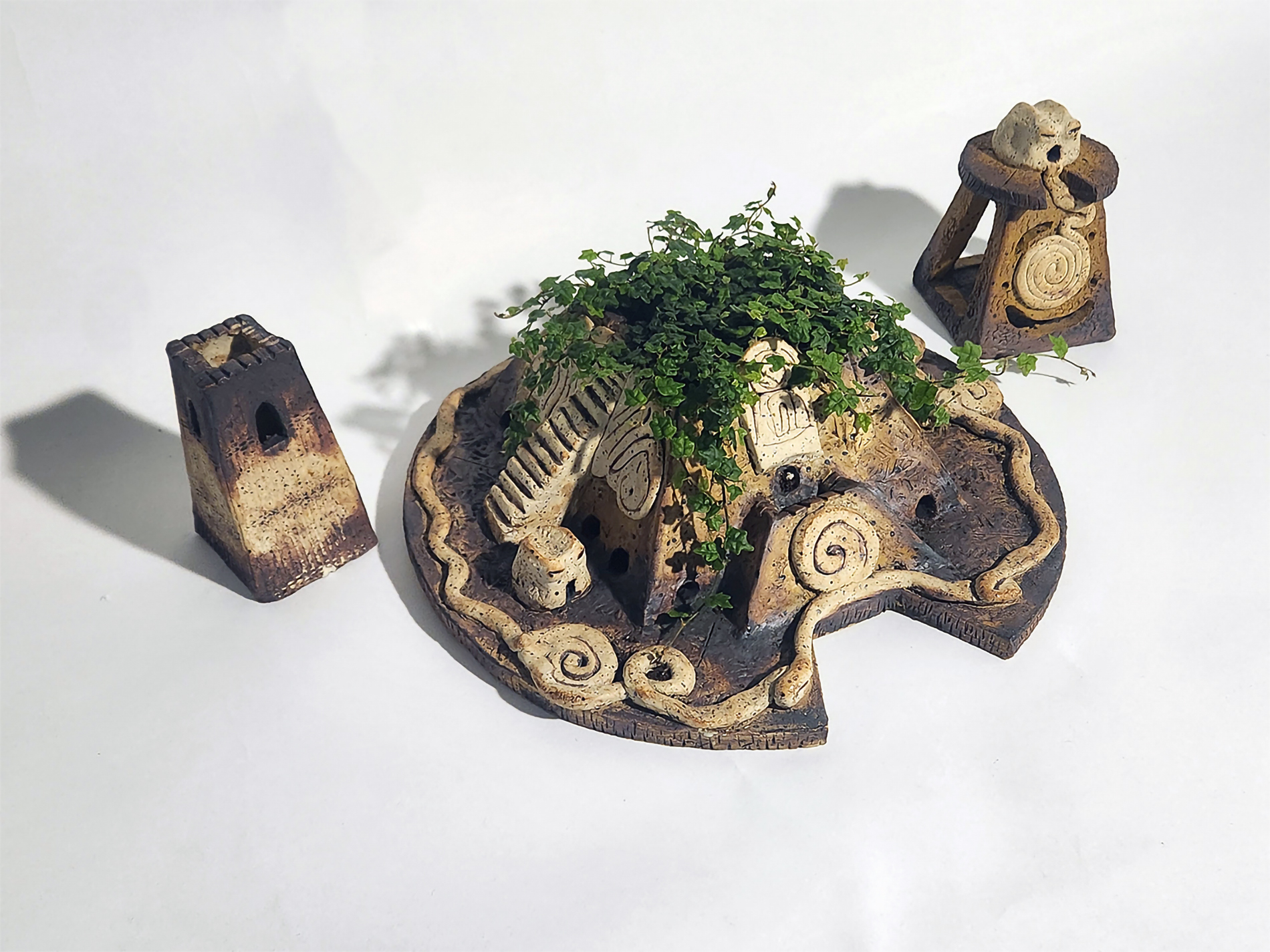 A ceramic structure with spiral patterns and greenery growing over it, surrounded by two separate tower structures on a white background, created by Kathleen Harton