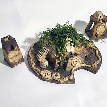 A ceramic structure with spiral patterns and greenery growing over it, surrounded by two separate tower structures on a white background, created by Kathleen Harton