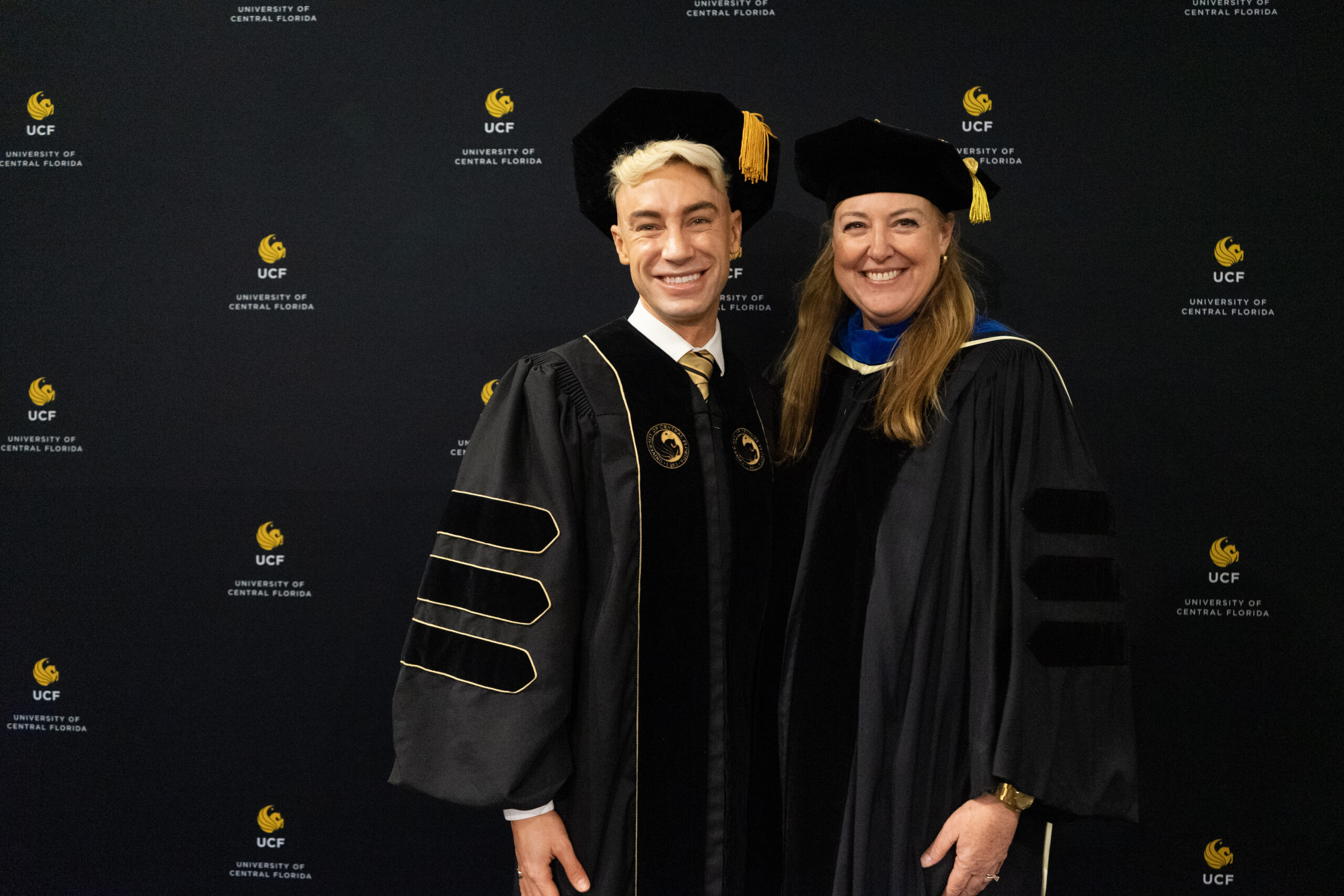 Two graduates in caps and gowns smile and wave during a graduation ceremony, surrounded by other graduates.