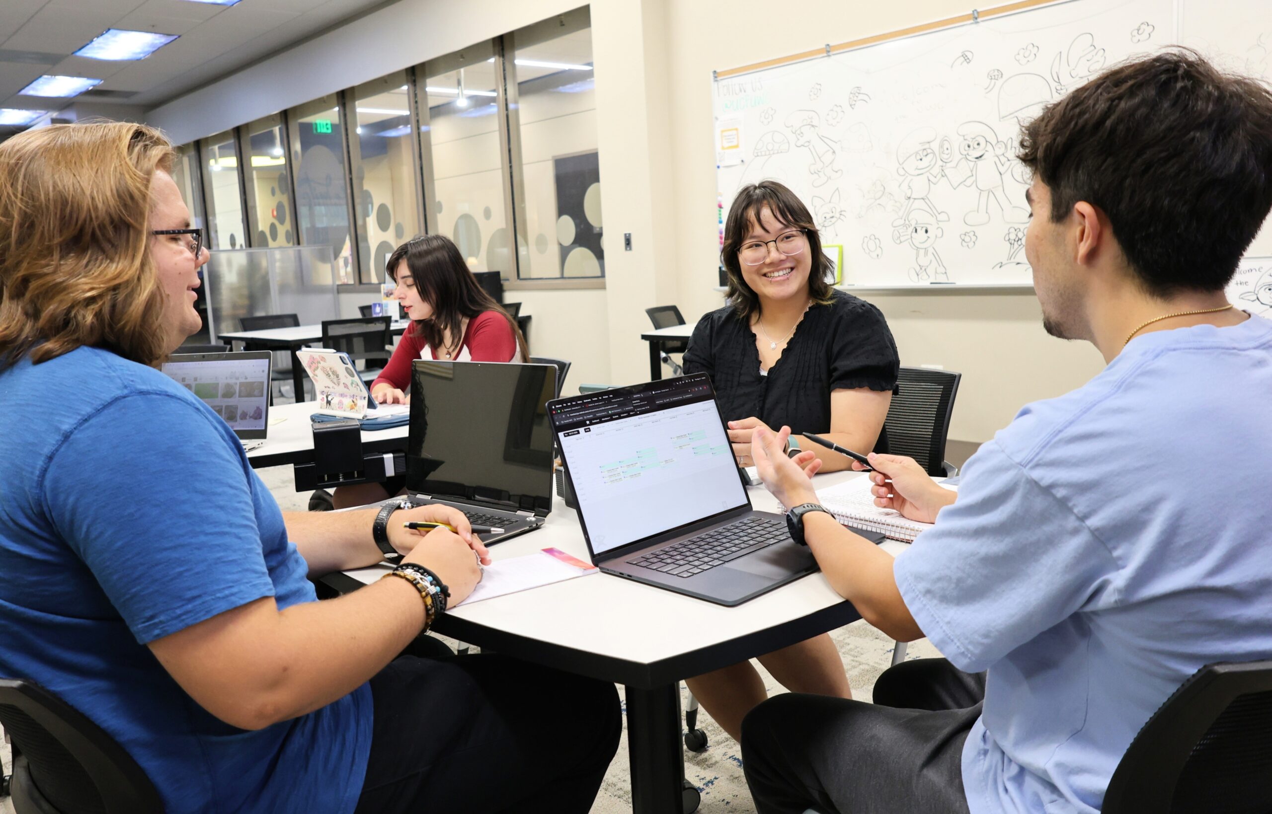 Four students collaborate around a table with laptops in a university writing center.