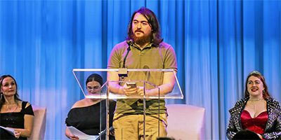 Student reads behind a podium while other students look on from the stage